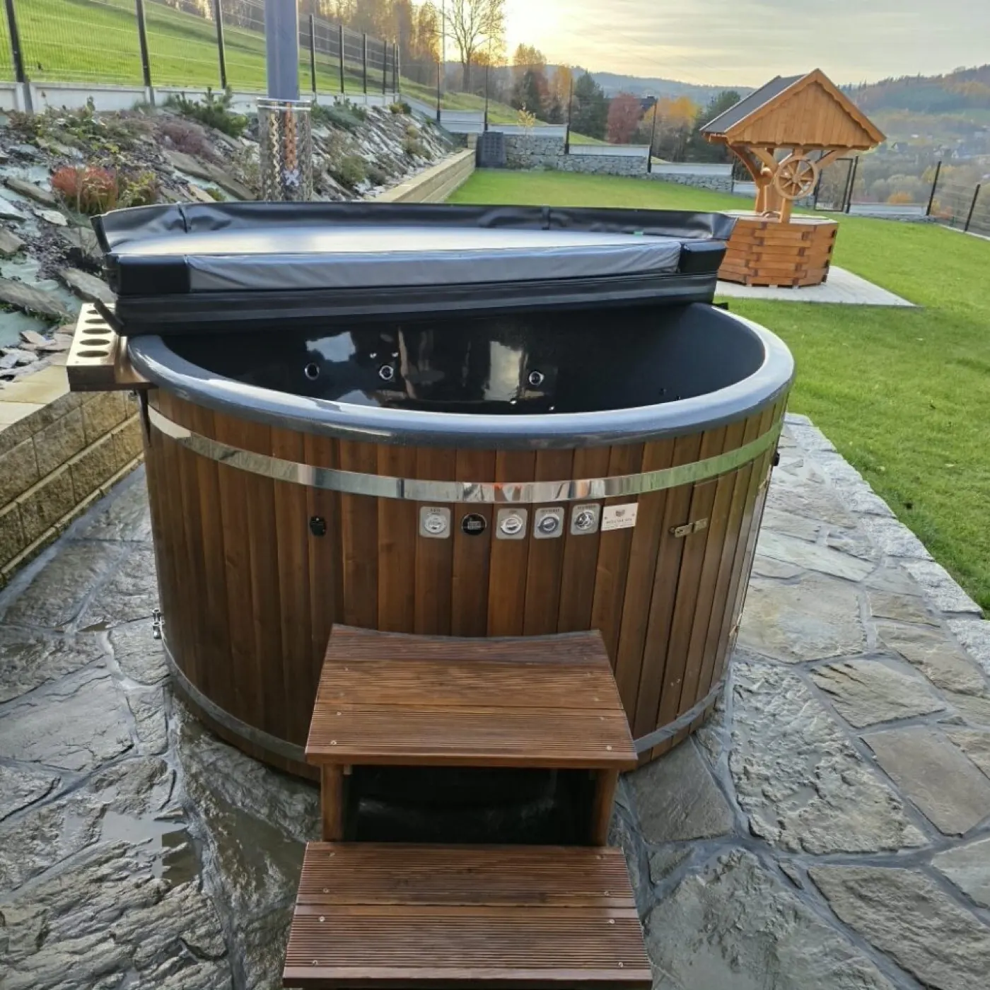 Okrągła wanna SPA ogrodowa na kamiennym tarasie o zachodzie słońca, z drewnianymi schodkami